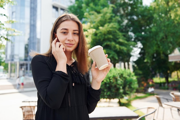 Per Telefon sprechen und Tasse trinken Glückliches junges Mädchen im schwarzen Rock im Freien in der Stadt in der Nähe von grünen Bäumen und gegen Geschäftsgebäude