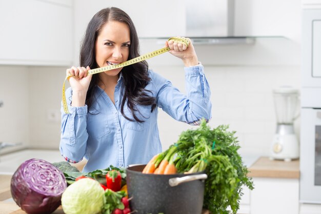 Perda de peso dieta e conceito de saúde - mulher jovem e bonita com vegetais frescos e fita métrica em sua cozinha.
