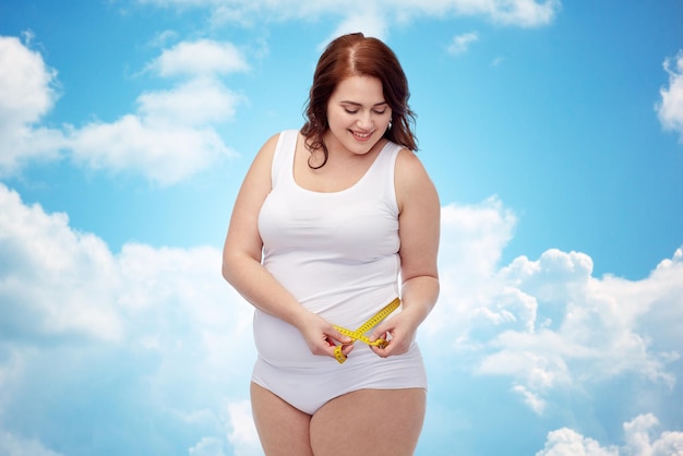 perda de peso, dieta, emagrecimento, tamanho e conceito de pessoas - feliz jovem mulher plus size em fita métrica de cueca sobre o fundo do céu azul e nuvens