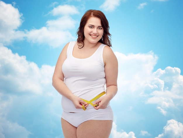 perda de peso, dieta, emagrecimento, tamanho e conceito de pessoas - feliz jovem mulher plus size em fita métrica de cueca sobre o fundo do céu azul e nuvens