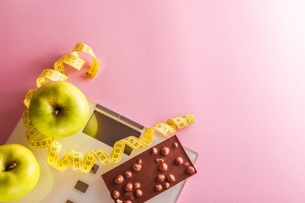 Perder peso concepto con escalas, cinta métrica, manzanas verdes y barra de chocolate sobre fondo rosa