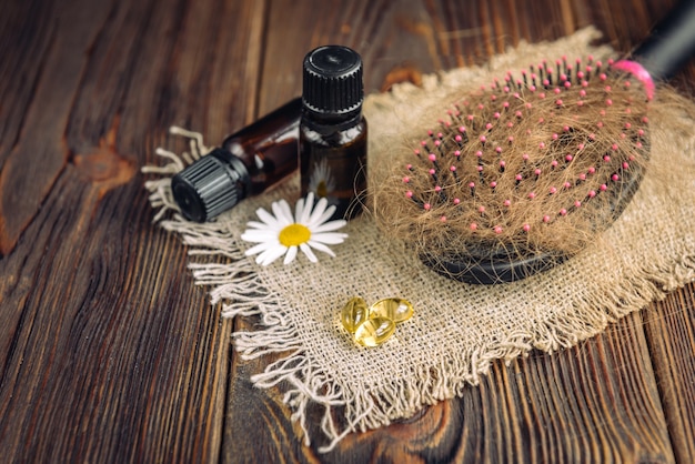 Perdida de cabello. Peinar con cabello y aceites esenciales, manzanilla y cápsulas de vitaminas sobre madera oscura.