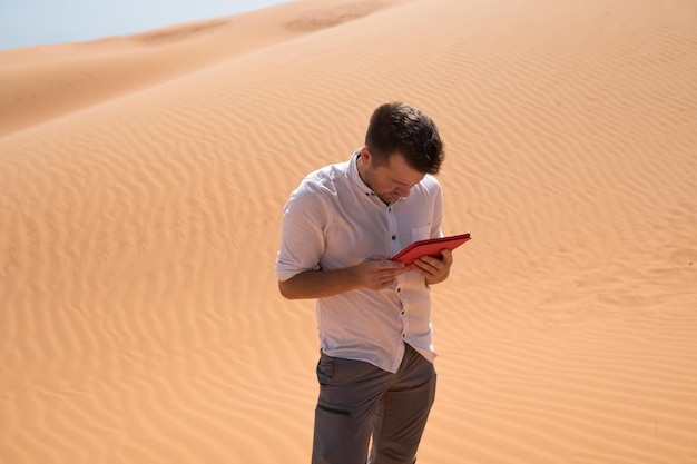 Perdido en el desierto Hombre caucásico sentado en la arena con tableta Está perdido y mirando en el mapa a dónde ir