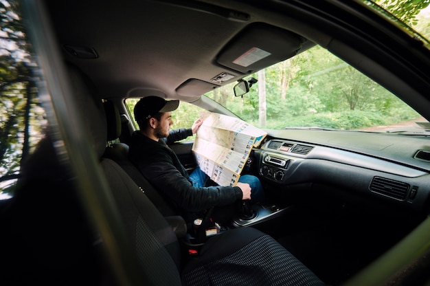 Perdido na floresta jovem piloto sentado no carro, mantendo o mapa e olhando de lado