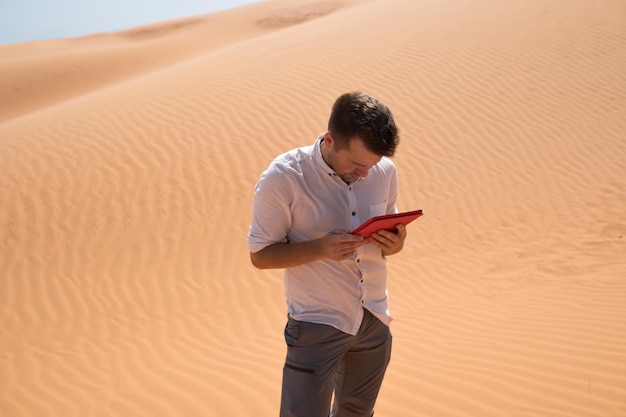 Perdido no deserto Homem caucasiano sentado na areia com tablet Ele está perdido e olhando no mapa para onde ir