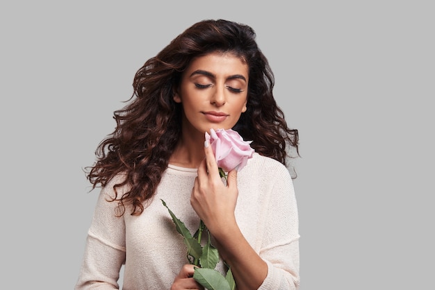 Perdido en sueños. Atractiva joven mujer sonriente manteniendo los ojos cerrados y sosteniendo una flor mientras está de pie contra el fondo gris