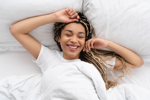 Perezosa, feliz, sonriente, joven mujer africana acostada en su cama blanca por la mañana.