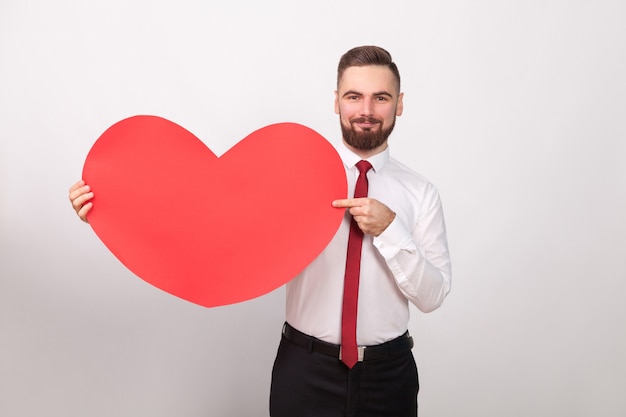 Perfecto hombre de negocios sonriendo, señalando con el dedo al gran corazón rojo. Interior, tiro del estudio, aislado en fondo gris