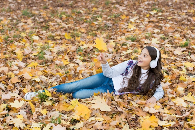 Perfekter Herbsttag des fröhlichen Mädchens Hörbuch oder Musik mit Kopfhörern in der Herbstsaison Parkspiel mit gelben gefallenen Ahornblättern entspannen