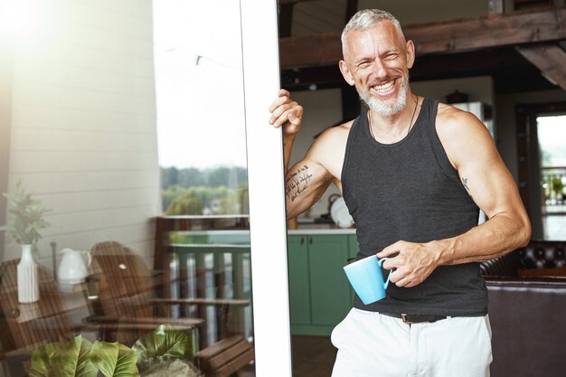 Perfekter Morgen schöner positiver kaukasischer Mann mittleren Alters mit Kaffee, der in der Nähe der Terrasse steht