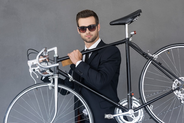 Perfekter Stadtverkehr. Selbstbewusster junger Geschäftsmann, der sein Fahrrad auf der Schulter trägt, während er vor grauem Hintergrund steht