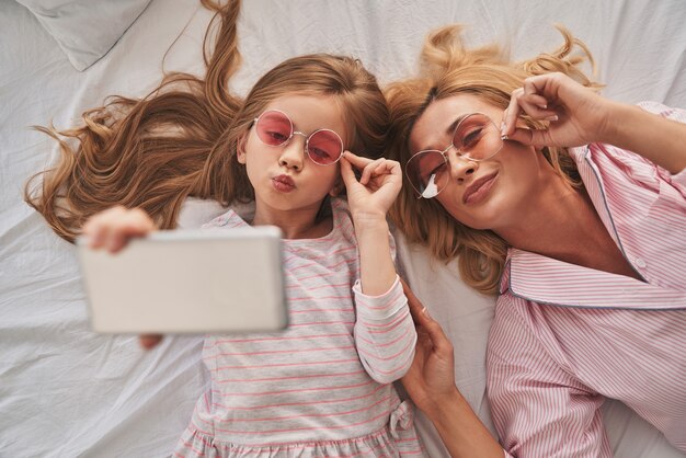 Perfektes Selfie. Draufsicht der jungen schönen Mutter und ihrer süßen Tochter, die Selfie mit Smartphone macht und lächelt, während sie zu Hause auf dem Bett liegt bed