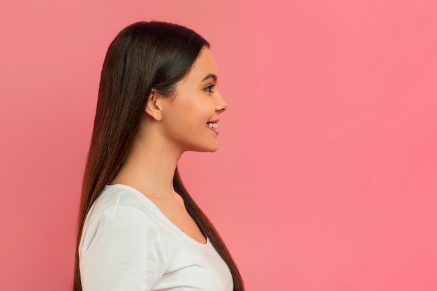 Foto perfil de una adolescente sonriente con el pelo largo de pie contra un fondo rosa