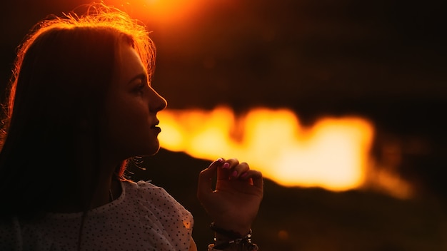 Perfil de una chica guapa que se lleva una mano a la cara con el telón de fondo de un lago al atardecer