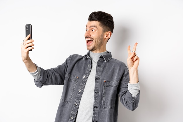 Perfil de jovem alegre tomando selfie no smartphone, mostrando o sinal V enquanto faz a foto no aplicativo móvel, em pé contra um fundo branco.