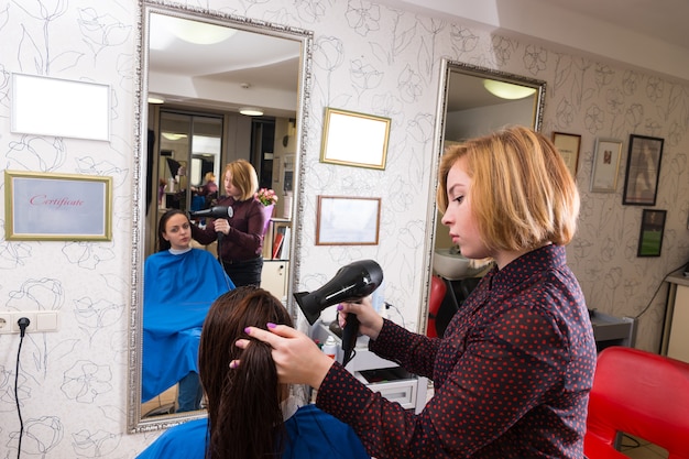 Foto perfil de jovem estilista loira secando o cabelo de uma cliente morena usando secador em salão de beleza com reflexo no espelho ao fundo