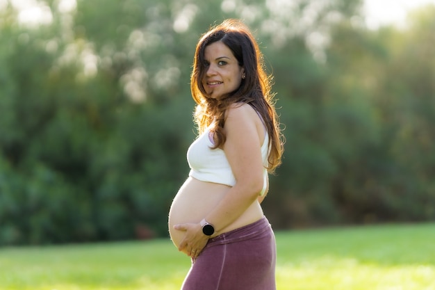 Foto perfil de uma mulher latina adulta grávida tocando seu abdômen olhando para a câmera, vestindo roupas esportivas confortáveis com o abdômen para cima em um parque com a luz do sol brilhando sobre ela