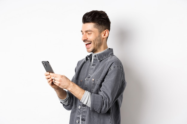 Perfil de hombre guapo charlando por teléfono, riendo y leyendo la pantalla del teléfono inteligente, de pie sobre fondo blanco.