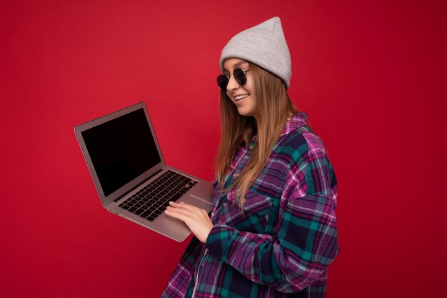 Perfil lateral hermosa mujer joven sosteniendo la computadora con ropa casual elegante, gafas de sol y sombrero