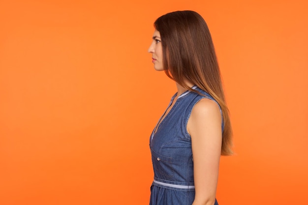 Foto perfil de una mujer hermosa con cabello largo y moreno y vestido de mezclilla de pie, mirando hacia un lado con rostro serio y tranquilo, expresión segura. foto de estudio aislada sobre fondo naranja, espacio de copia