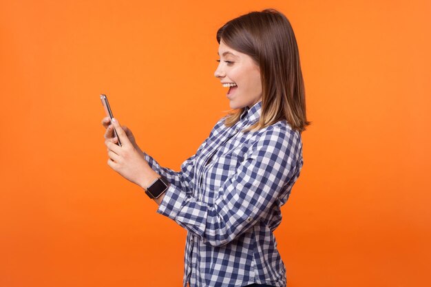 Perfil de mujer morena hermosa asombrada con una sonrisa encantadora en camisa a cuadros leyendo un mensaje en el teléfono celular gratamente sorprendida por buenas noticias en un estudio interior aislado en un fondo naranja