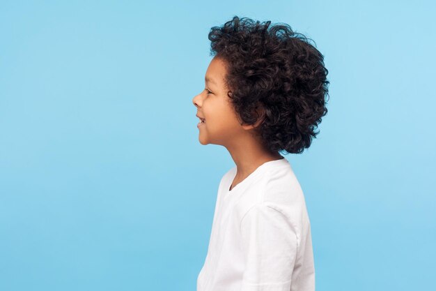 Perfil de niño feliz con el pelo rizado en camiseta sonriendo despreocupado y mirando al espacio de copia lateral, niño sano y alegre con emociones positivas. Foto de estudio interior aislado sobre fondo azul.