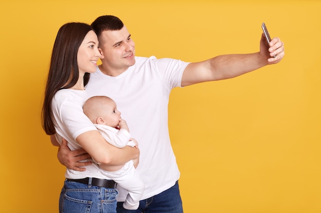 Perfil de pareja feliz tomando selfie con bebé en manos, mirando con expresiones faciales alegres en la parte delantera del dispositivo, pareja con hija pequeña aislada sobre pared amarilla.