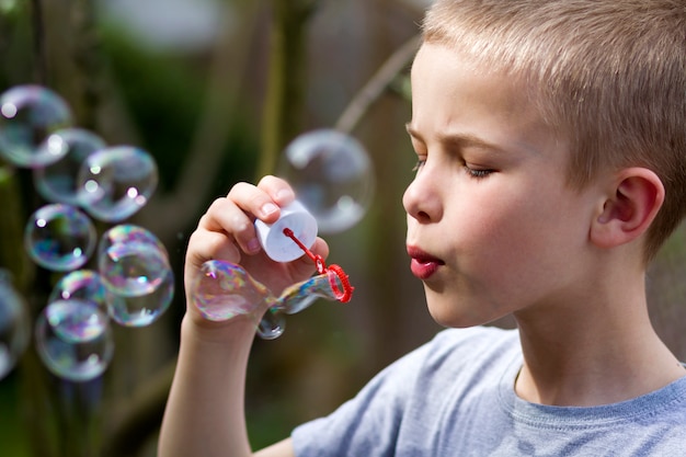 Perfil del pequeño muchacho rubio hermoso lindo del niño con la expresión seria divertida que sopla burbujas de jabón transparentes coloridas al aire libre en escena verde borrosa del verano. Alegría del concepto de infancia descuidada.