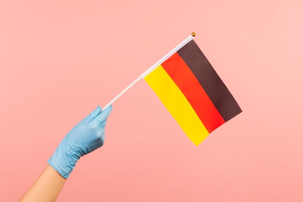 Perfil vista lateral closeup de mão humana em luvas cirúrgicas azuis segurando a bandeira da Alemanha.