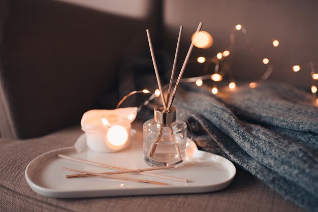 Perfume casero líquido en botella de vidrio con palos de bambú de madera y vela encendida en bandeja de cerámica blanca, cárdigan de lana tejido en una acogedora silla en la habitación de cerca. Buenos días. Ambiente confortable y higiénico.