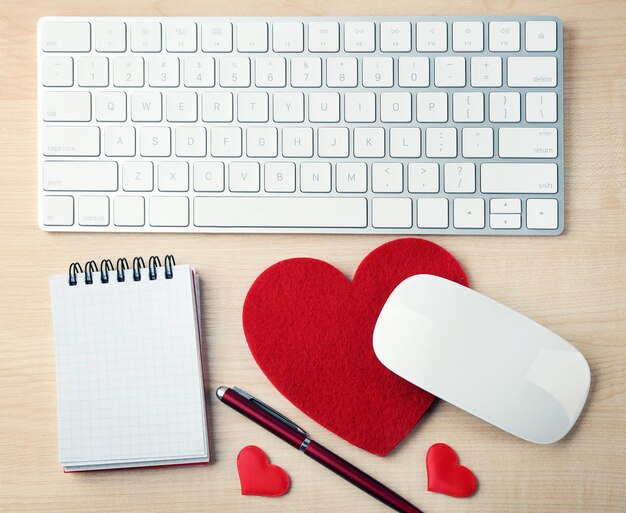 Periféricos de computadora con bolígrafo de corazones rojos y cuaderno en mesa de madera clara