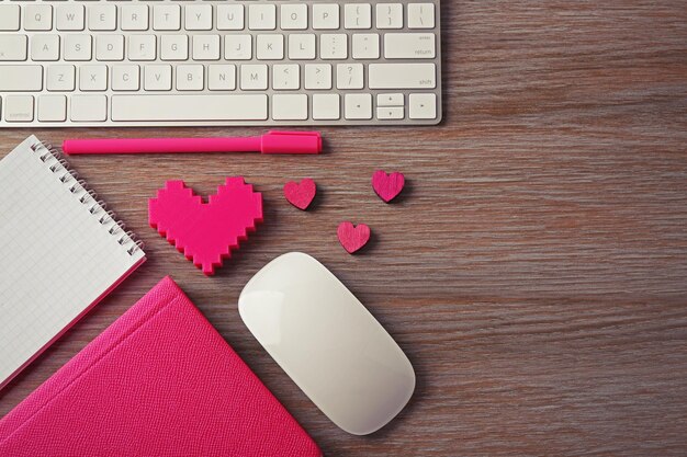 Periféricos de computadora con bolígrafo de corazones rosas y cuadernos en mesa de madera