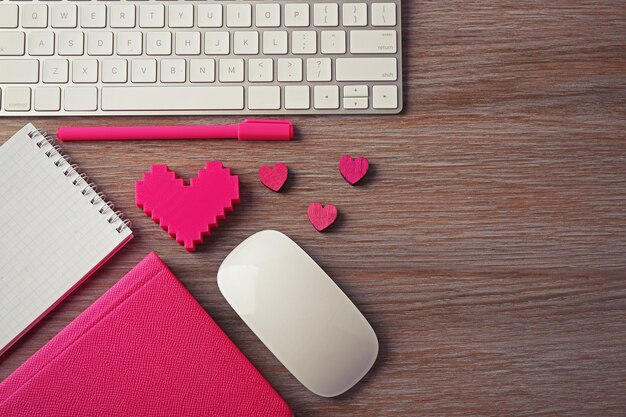 Periféricos de computadora con bolígrafo de corazones rosas y cuadernos en mesa de madera