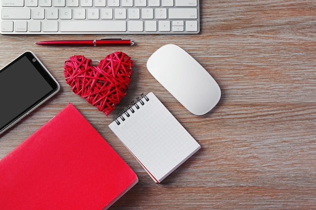 Periféricos de computadora con cuadernos de corazón de mimbre rojo y teléfono móvil en mesa de madera