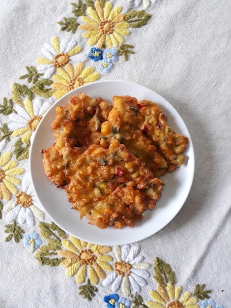 Perkedel Jagung o Bakwan Jagung Buñuelos de maíz tradicionales indonesios servidos en un plato blanco con un mantel bordado floral en el fondo