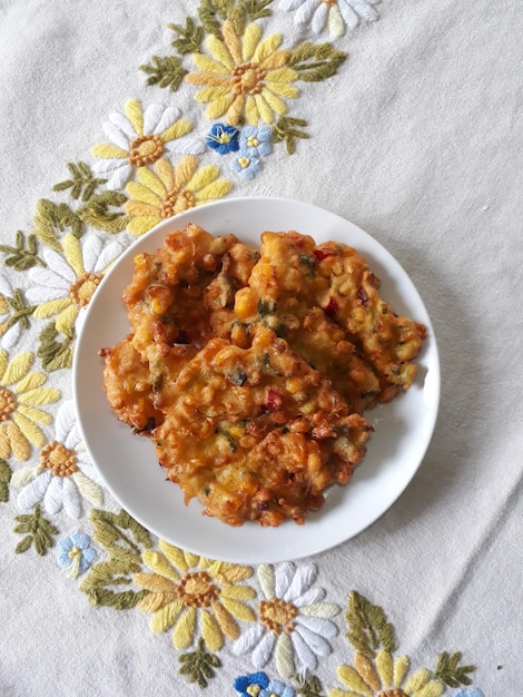 Perkedel Jagung o Bakwan Jagung Buñuelos de maíz tradicionales indonesios servidos en un plato blanco con un mantel bordado floral en el fondo