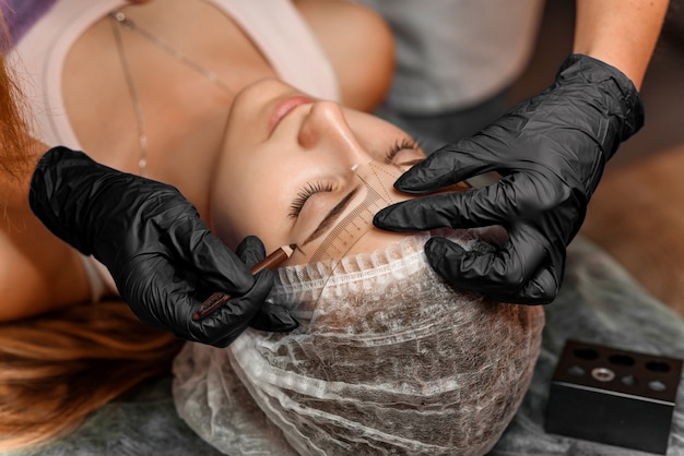 Permanentes Brauenmake-up im Schönheitssalon, Nahaufnahme. Professionelle Kosmetikerin markiert die Länge der Augenbrauen mit einem Bleistift und einem speziellen Lineal zum Messen der Augenbrauen. Kosmetologische Behandlung.