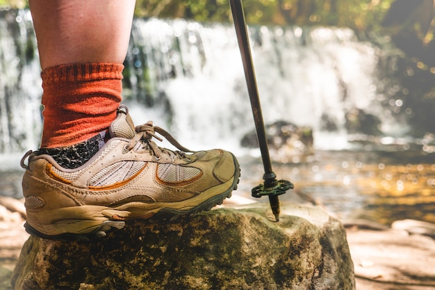 Foto perna de mulher com botas de caminhada e bengala em uma rocha com uma cachoeira natural atrás