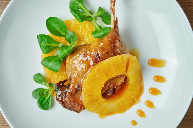 Perna de pato apetitoso confit com abacaxi e espinafre em um prato branco sobre uma mesa de madeira. restaurante que serve do chef