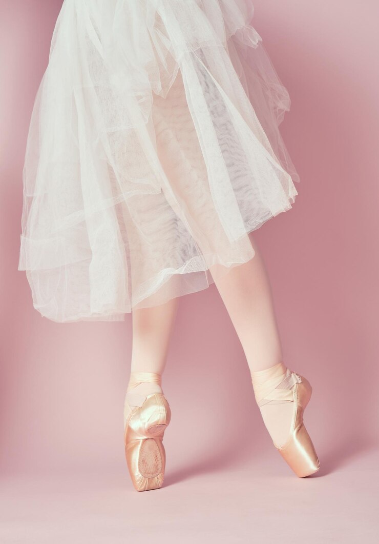Foto pernas de bailarina clássica vestindo meia-calça e saia branca na ponta dos pés com sapatilhas de balé no fundo rosa