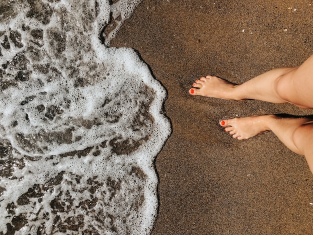 pernas de mulher com os pés descalços nas ondas de espuma do mar na praia de areia dia de verão. vista de cima acima dos pés femininos