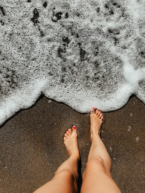 pernas de mulher com os pés descalços nas ondas de espuma do mar na praia de areia dia de verão. vista de cima acima dos pés femininos