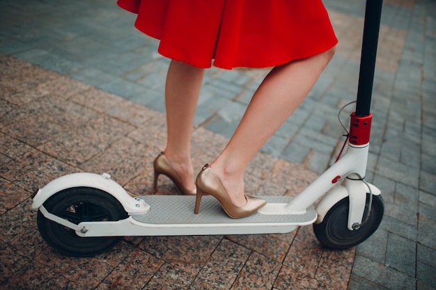 Pernas de mulher com scooter elétrico de vestido vermelho na cidade