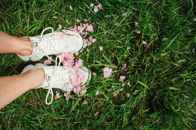 Pernas de mulher de tênis branco na grama verde com flores de sakura florescendo rosa copiam o espaço