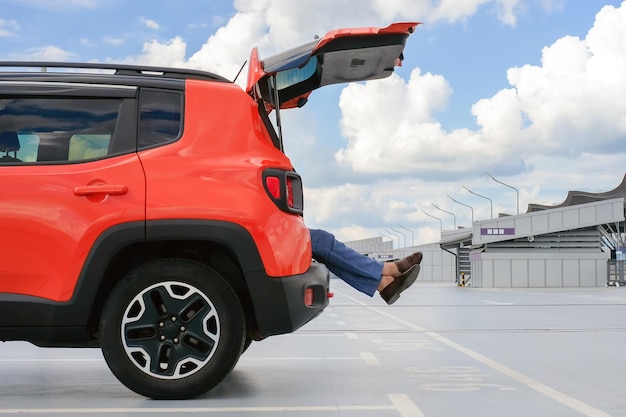 Pernas de um homem do porta-malas de um carro vermelho em um estacionamento do aeroporto visão lateral do conceito de férias