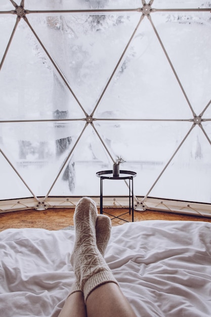 Pernas de uma mulher em meias de lã em uma cama no contexto de uma floresta coberta de neve em um acampamento sob uma cúpula