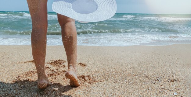 Pernas femininas com um chapéu branco na mão no fundo da paisagem. Conceito de férias e turismo na praia. Espaço para texto.