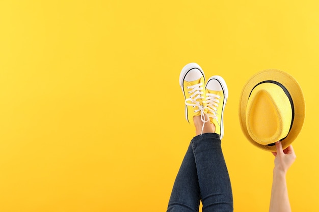 Pernas femininas em jeans e tênis e mão feminina segurando o chapéu em fundo amarelo