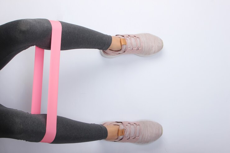 Foto pernas femininas em leggings esportivos e tênis, fazendo exercícios com elástico de fitness na superfície branca. vista do topo