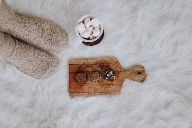 Pernas femininas em meias quentes perto de uma xícara de chocolate quente com marshmallow e bolo de brownies em bandeja de madeira